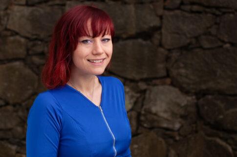 Portrait of a smiling woman with red hair wearing a blue top against a stone wall